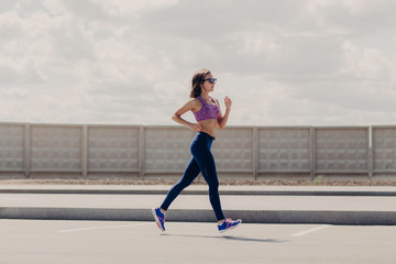 Outdoor shot of athletic female jogger with slim body runs on road, prepares for running marathon,...