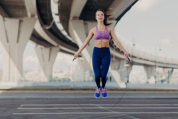 Fototapeta na wymiar Fit girl in good shape jumps on skipping rope, wears purple top and leggings, likes exercices outdoor, cares of her body. Athletic young woman has workout in open air. People and fitness concept