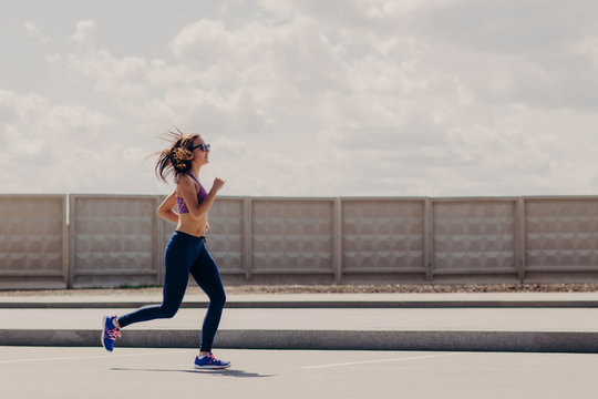 Horizontal shot of sportive woman joggs in open air, enjoys sport activities, wears sport clothes, concentrated into distance, has running workout. Female runner motivates yuo to go in for sport