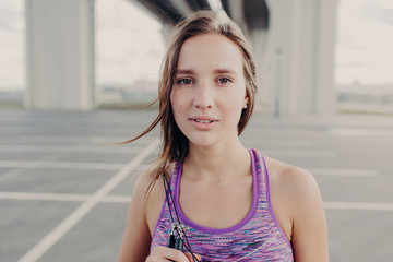 Close up portrait of sporty woman with healthy clean skin, serious expression, dressed in purple top, uses flipping rope for gymnastics, poses outdoor, breathes fresh air, has workout alone.