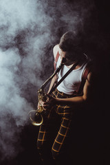 Fototapeta na wymiar high angle view of stylish young musician playing saxophone in smoke on black