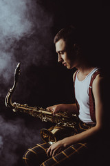 Obraz na płótnie Canvas side view of stylish young jazzman sitting and holding saxophone in smoke on black