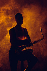 silhouette of young musician sitting on stool with saxophone in smoke