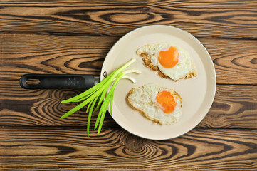 Two fried eggs in metal beige pan and green fresh onion on old wooden brown table. Top view