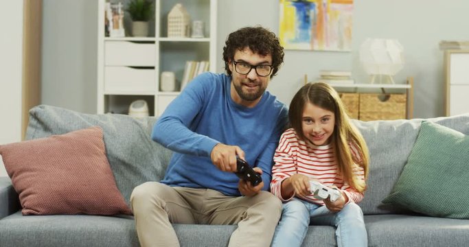 Caucasian happy small pretty girl playing videogames with her handsome father with controls in their hands, then daughter screeming joyfully because she winning. Indoors.