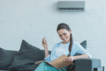 woman turning on air conditioner with remote control while reading book on sofa