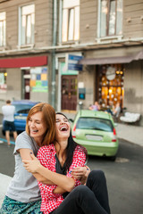 Two young girlfriends hugging, laughting and sitting near road. one girl with long brunette hair in red plaid shirt, another redhead girl in gray shirt and blue skirt. concept of sincere friendship