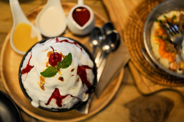 Bingsu with sweet red jam on black cup and wooden tray with syrup place on table,A red cherry and mint put on white milk Bingsu,Selective focus