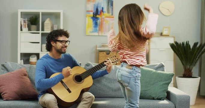Happy father playing guitar and sitting on the couch while his cute daughter singing and dancing. Indoors.