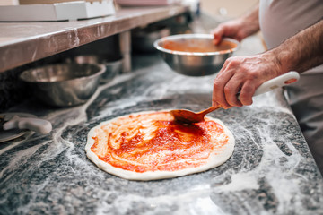 Raw pizza, preparation in traditional style. Adding tomato sauce on a pizza dough. - obrazy, fototapety, plakaty