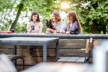 Young girlfriends having fun together sitting with dog at the outdoor park cafe
