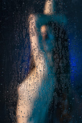 Blurred silhouette of a beautiful slender naked girl standing behind glass covered with drops of...
