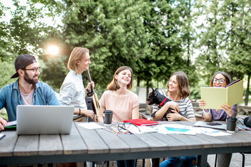 Young students having fun together meeting a friend while sitting with dog at the table outdoors