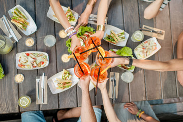 Friends clinking glasses with cocktail drinks, top view on the table full of snacks outdoors - obrazy, fototapety, plakaty