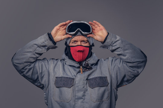 Portrait of a snowboarder dressed in a full protective gear for extream snowboarding puts on glasses while standing at a studio. Isolated on gray background.