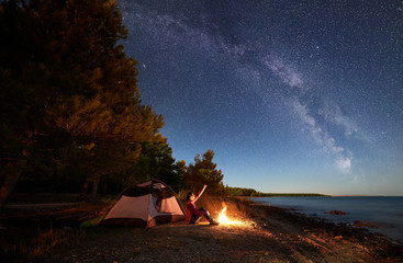 Night camping at sea shore. Female backpacker sitting in front of tourist tent at campfire near forest, pointing to sky full of stars and Milky way, enjoying beautiful view of clear blue water