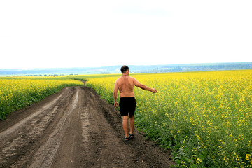 A young man walks along a dirt road, along a field of yellow flowers.