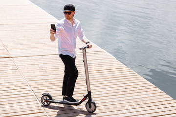 Modern man in stylish outfit using his smartphone while standing at wooden pier with electric scooter