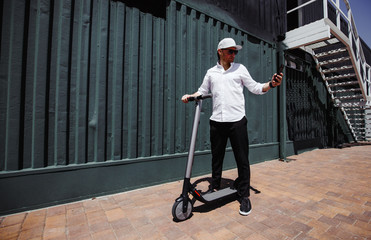 Modern man in stylish outfit using his smartphone while standing at street with electric scooter