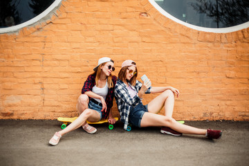 Two young happy girl friends in hipster outfit sitting on longboards and making selfie on phone