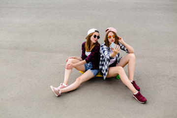 Two young happy girl friends in hipster outfit sitting together on longboard and making selfie on...