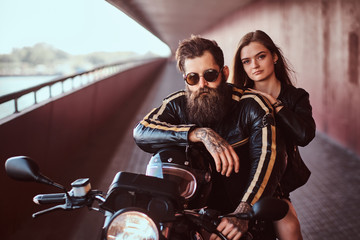 Fototapeta na wymiar Brutal bearded biker in a black leather jacket with sunglasses and sensual brunette girl sitting together on a custom-made retro motorcycle on footway under a bridge.