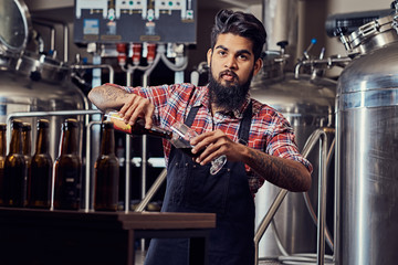 Stylish full bearded Indian man in a fleece shirt and apron pours beer in a glass for quality control, standing behind the counter in a brewery.