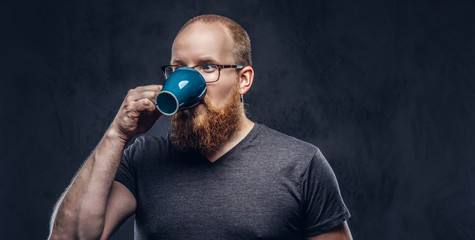 Close up portrait of a redhead bearded male drinks coffee wearing glasses dressed in a gray t-shirt, isolated over a dark textured background.