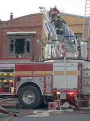 Two firefighters descending ladder on fire trick after putting out a blaze in a two story building on an urban commercial street. Broken window and charred interior visible in background
