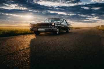 Black retro vintage muscle car is parked at countryside asphalt road at golden sunset - 213989554