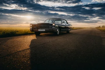 Fotobehang Zwarte retro vintage muscle car staat geparkeerd op de asfaltweg op het platteland bij gouden zonsondergang © Ivan Kurmyshov