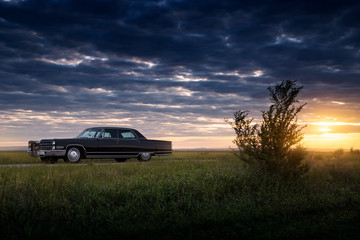 Black retro vintage muscle car is parked at countryside asphalt road at golden sunset