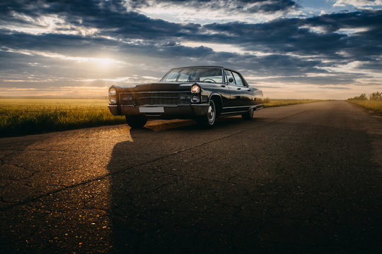 Black retro vintage muscle car is parked at countryside asphalt road at golden sunset