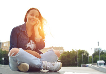 A beautiful girl in a jeans jacket, sitting on the steps on a city street, uses her smartphone. A woman communicates through the Internet.