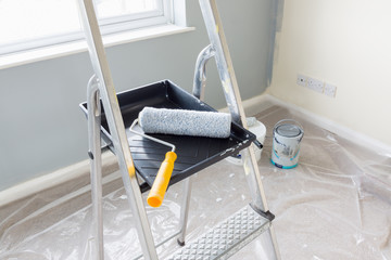 Painting and decorating with a roller and tray on a set of metal step ladders with cans of paint and a dust sheet in the background - obrazy, fototapety, plakaty