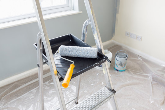 Painting and decorating with a roller and tray on a set of metal step ladders with cans of paint and a dust sheet in the background