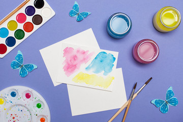 A view from above of the artist's creative accessories: brushes, paints, a palette, a picture and blue paper butterflies. Place for text, workpiece, back violet background. flat lay