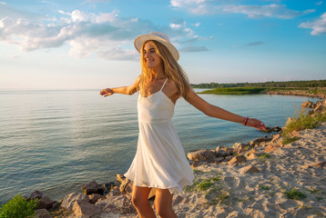 Beautiful happy young woman in white summer satin dress and wearing a hat whirls at the sea...