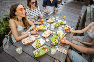 Young friends having fun sitting together with tasty snacks and drinks during the evening lights at...