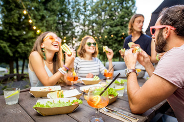 Young friends having fun sitting together with tasty snacks and drinks during the evening lights at the park cafe outdoors