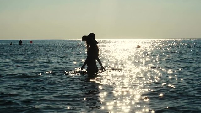Beautiful young woman walking on the water in sunlight, silhouette of girl in fashionable hat walking on the sea water. Sun flare, slow motion