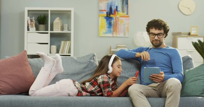 Portrait shot of the handsome father sitting on tthe sofa in the living room and using tablet device while his daughter lying beside and listening to the music on the smartphone. Indoors.