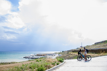 Fototapeta premium two teenage girls on bicycles on a concrete path to the sea in summer. Water landscape with clouds on the horizon. Sea nature of tranquility. The weather is changing.