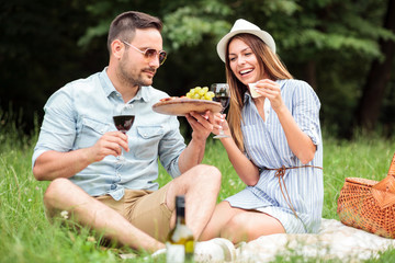 Fototapeta premium Happy young couple celebrating anniversary or birthday by having a romantic picnic in park. Eating cheese, grapes and drinking red wine. Love and dating, romance, lifestyle concept