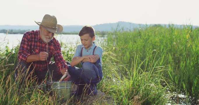 Senior fisherman in a hat with his grandson sitting on the shore of the lake and putting fish in the backet after angling. Outdoor.