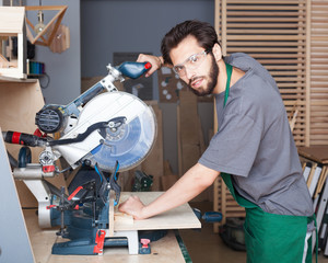 Young hipster man in black overalls by profession carpenter builder saws with a circular saw a wooden board on a wooden table in the workshop