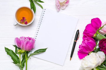 Empty notebook with place for text, pen, a cup of tea and peonies flowers on a white wooden background. Mockup for your design. View from above