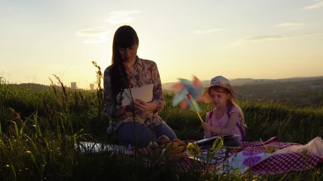The family sits on a hill on a picnic in the rays of the setting sun. A young mother draws with a pencil, a small daughter holds a rotating pinwheel in her hand.