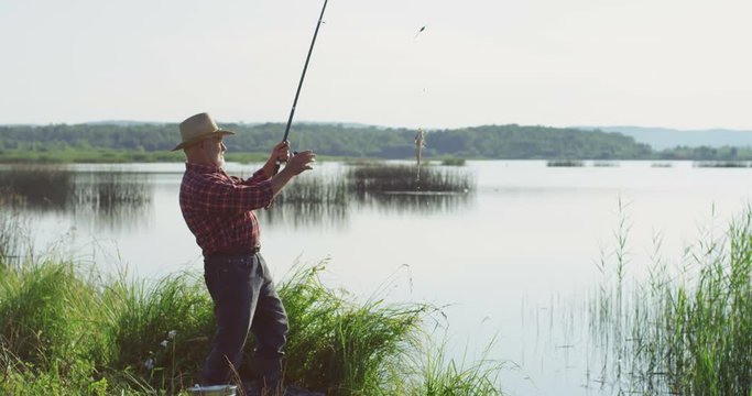 Old gray haired Caucasian fisherman in a hat catching a fish on a rod and getting it out on the lake. Outdoors.