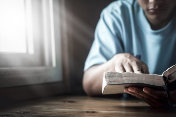 Young man reading Holy Bible next to the window at home.